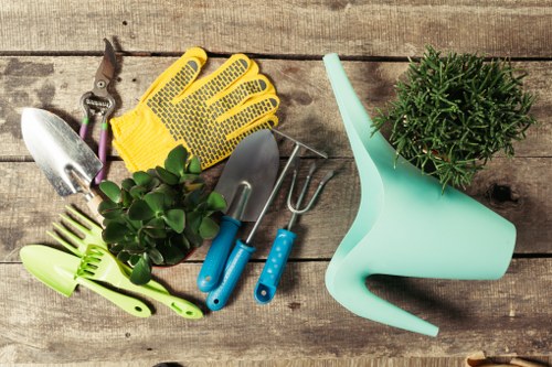 A flat-lay image of various gardening tools and accessories arranged on a weathered wooden surface, typical of outdoor gardening work in the Hendon area. Included are a pair of yellow gardening gloves, a small silver trowel, a hand fork, and a cultivator, all positioned around a small potted shrub with dense green foliage. A light blue plastic watering can with a long spout is placed nearby, alongside a green-handled plant spray. The tools are well-organized, indicating preparedness for garden maintenance tasks such as planting, watering, or trimming. The wooden surface provides a rustic background that complements the garden scene, reflecting typical outdoor spaces found in Hendon households. The natural tones of the wood contrast with the vibrant green of the plants and the bright colours of the tools, suggesting an active gardening environment suitable for lawn care, shrub trimming, and outdoor beautification services offered by Landscapers Hendon. The lighting indicates day-time conditions, with soft natural light enhancing the clarity and detail of each item, reinforcing the connection between outdoor maintenance and professional garden care services in the local area.