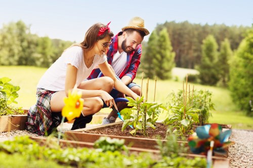 In a sunny outdoor garden, a man and a young girl are engaged in planting and caring for a vegetable patch. The girl, wearing a white T-shirt with a pink headband, is crouched down and using a small gardening tool to tend to leafy green plants. The man, dressed in a plaid shirt, white T-shirt, and wearing a straw hat, is nearby, assisting and observing the gardening activities. The vegetable bed is bordered by wooden planks and contains young, healthy plants with broad green leaves. Surrounding the garden are neat, grassy areas and a backdrop of tall, lush trees under a clear sky, indicating spring or summer weather. The setting appears to be a well-maintained front or backyard space, typical of a residential garden in Hendon or North London, with natural lighting highlighting the vibrant greenery and natural soil surface. The scene reflects outdoor gardening and lawn care activities, consistent with professional landscaping and gardening services offered by Landscapers Hendon, with a focus on family-friendly outdoor spaces and vegetable cultivation.