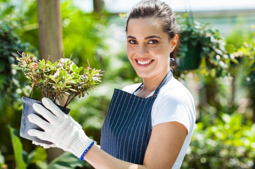 A young woman with dark hair tied back, wearing a navy apron and white gardening gloves, is standing outdoors in a well-maintained garden. She is smiling at the camera while holding a small potted shrub with green and pink-tinged leaves. The background features a lush, green environment with various plants, including bushes, flowering plants, and a hedge, indicating a healthy and vibrant garden setting. Behind her, there are wooden garden structures and a hint of sunlight filtering through the foliage, suggesting a clear day. The scene showcases a neat lawn and well-kept plant borders, with a natural and inviting outdoor atmosphere typical of residential gardens in Hendon or similar areas, with landscaping and gardening services by Landscapers Hendon supporting the garden's aesthetic and health.