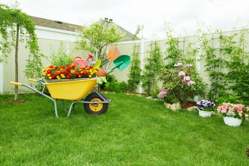 A well-maintained backyard garden featuring a lush, green lawn with dense, evenly cut grass in the foreground. To the right, there is a flower bed bordered by red brick edging, filled with blooming pink and purple flowers, alongside a white pot containing pink flowers. In the middle background, a small shrub with green foliage is visible, surrounded by climbing plants growing along a white-painted wooden fence. A yellow garden wheelbarrow, parked on the lawn, is loaded with colorful plastic gardening tools such as watering cans and small shovels, indicating ongoing gardening activities. The scene is lit by natural daylight under a partly cloudy sky, creating a bright and inviting outdoor environment. This garden layout showcases a combination of neatly kept lawn and flower borders, exemplifying professional gardening and outdoor space maintenance that a company like Landscapers Hendon might undertake for local residential properties in Hendon and the surrounding NW postcode area.