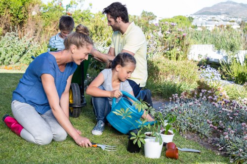 In a well-maintained suburban garden, a family of four is engaged in gardening activities on a sunny day. The foreground features a lush, green lawn with evenly cut grass, and a variety of flower beds bordered with natural stones and rich soil, exhibiting colorful blossoms and healthy plants. A woman in a blue top and grey trousers is kneeling on the grass, tending to a small plant, while a young girl in denim jeans and a blue backpack is actively watering another plant with a garden spray. Behind them, an adult man in a light shirt and a boy in a striped shirt are engaged in conversation, with the man holding garden tools or equipment, suggesting outdoor maintenance or planting. The background reveals more garden vegetation including shrubs, flowering bushes, and taller trees, with a glimpse of a distant town or cityscape over the horizon. The scene is illuminated by natural daylight under clear weather, emphasizing the vibrant natural tones of greens, browns, and the blooming colors, fitting with professional garden and landscaping services offered by Landscapers Hendon.