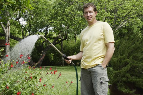 A man stands in a lush, well-maintained garden in Hendon, holding a garden hose and watering nearby flowering plants with small red blossoms. Behind him, a neatly trimmed hedge runs parallel to a vibrant green lawn, with several trees and shrubs providing a layered landscape. The garden features a gravel pathway and patches of soil bordering flower beds, all under a partly cloudy sky with natural sunlight illuminating the scene. The environment appears tidy and cared for, reflecting professional gardening and outdoor maintenance services typical for gardens in the Hendon area, Essex, or surrounding London suburbs. This image showcases a peaceful outdoor space, suitable for lawn care, hedge trimming, and general garden landscaping, highlighting the routine work involved in maintaining a healthy, attractive garden as part of a comprehensive gardening service. A subtle emphasis on natural colours like green, red, and earthy tones underscores the quality and care typical of landscapers serving the local community.