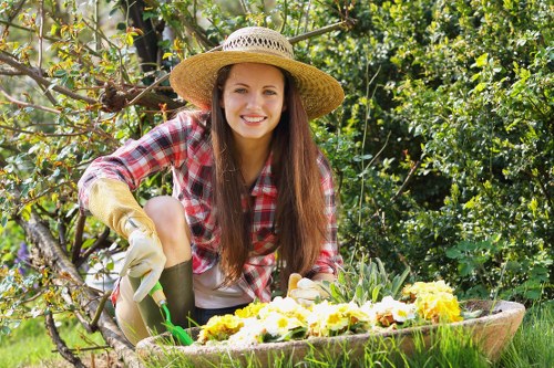 A young woman with long brown hair smiling while working in a garden, wearing a straw hat, a red and white checkered shirt, and gardening gloves. She is kneeling beside a round, green garden bed filled with blooming yellow and purple flowers, with a background of lush green bushes, trees, and foliage. The garden appears well-maintained with healthy plants and a natural, outdoor setting, typical of a landscaped yard in Hendon, London. The scene is brightly lit, suggesting a sunny day, and the overall environment reflects a tidy, cultivated outdoor space suitable for gardening services focused on plant care and landscape maintenance.