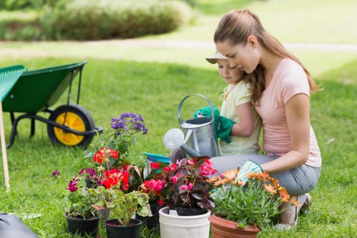 A woman and a young girl are kneeling on a well-maintained grassy lawn in a garden, tending to a variety of potted flowering plants, including colorful petunias and other blooms, with a focus on vibrant reds, pinks, and purples. The garden features a lush, green lawn bordered by a curved hedge in the background, with a paved pathway or patio area partially visible. Nearby, a green garden wheelbarrow with yellow wheels is positioned on the grass, suggesting ongoing gardening activity. The garden appears to be part of a residential outdoor space, well-kept and suitable for family gardening and outdoor leisure, indicative of professional gardening services in Hendon aiming to maintain attractive, eco-friendly outdoor environments. Overcast weather provides soft, diffuse lighting, enhancing the natural tones of the plants and surroundings. The scene captures a moment of gardening care, emphasizing the importance of outdoor maintenance and landscape upkeep, consistent with the services offered by Landscapers Hendon across the local area.