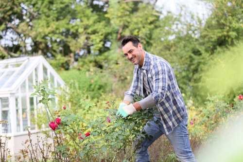 A man is working outdoors in a well-maintained garden, tending to flowering rose bushes with pink blooms. He is wearing a casual plaid shirt and jeans, using gardening gloves, and smiling as he actively prunes or cultivates the plants. The garden features a neatly manicured lawn in the foreground with lush green grass, bordered by flower beds containing a mix of vibrant and natural plant colours. In the background, there are mature trees with dense foliage and a small greenhouse structure with transparent panels, suggesting a well-organised outdoor space suitable for gardening services in the Hendon area. The scene is illuminated by natural daylight under a clear sky, indicating favorable weather conditions for outdoor garden maintenance. This setting illustrates typical features of a landscaped residential garden that local gardening and landscaping companies like Landscapers Hendon might service, highlighting their expertise in garden care and plant management.