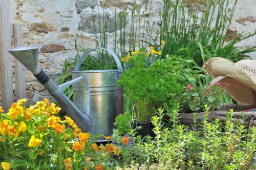 The image depicts a small, neatly maintained garden corner with a variety of flowering plants and greenery. In the foreground, there are vibrant yellow and orange flowering plants, likely suitable for garden beds or border planting. Behind these, taller green plants with slender leaves and some tiny pink flowers add height and diversity to the plant arrangement. A weathered, metal watering can is prominently positioned in the middle, indicating active garden care. To the right of the watering can, there is a lush shrub with finely divided green foliage, contributing to the garden's natural texture. A wide-brimmed straw hat rests on a surface or plant nearby, suggesting active outdoor gardening. The backdrop features a rustic stone wall, enhancing the outdoor garden environment with natural tones and textures. The lighting appears natural, possibly from a bright, sunny day, with slight shadows cast across the plants. The scene reflects a well-tended outdoor space, typical of residential gardens in the Hendon area, where gardening services by Landscapers Hendon might focus on plant care, lawn maintenance, and garden enhancement, creating a charming, inviting garden environment.