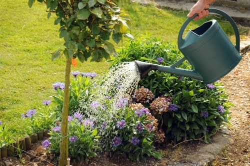 In a well-maintained garden, a person is watering a flower bed using a green watering can held in their right hand. The flower bed features a variety of blooming plants, including light purple and pink flowers, bordered by small wooden edging. Behind the flower bed, there is a lush, green hedge and a small tree with a leafy canopy. The garden's grassy lawn is visible in the background, with vibrant green grass extending across the scene. The weather appears to be sunny, casting natural light over the garden. The overall setting showcases a tidy outdoor space that may be part of a residential property in Hendon or surrounding areas, ideal for services offered by Landscapers Hendon in garden landscaping and maintenance.