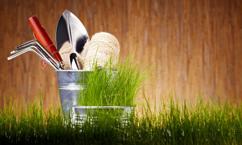 A close-up of a small garden scene featuring two metal containers placed on lush, green grass, with one container holding gardening tools including a trowel, a cultivator, a pruning saw, and a ball of twine. The tools have metal and wooden handles, contrasting against the bright green grass, which appears healthy and dense. The background consists of a warm, wooden surface, suggesting a garden or outdoor shed wall, providing a natural and inviting setting. This imagery highlights essential gardening tools and materials that a professional landscaper or gardener in Hendon might use for outdoor maintenance, landscaping, or planting projects, aligning with gardening services offered by Landscapers Hendon. The scene is captured in natural light, emphasizing the vivid greenery and the clean, shiny surfaces of the gardening tools, suitable for supporting content about garden care, tool availability, or outdoor enhancements within the Hendon area, postcode NW4, in a professional and factual tone.