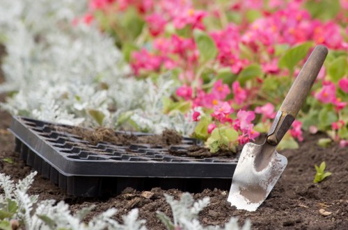 A gardening scene in a landscaped outdoor area, featuring a close-up view of a freshly prepared flower bed with dark, moist soil. A small metal garden trowel with a wooden handle is inserted into the soil, indicating recent planting activity. Surrounding the bed, there are delicate, low-growing plants with silvery leaves, and in the background, vibrant pink and white flowering plants add colour to the scene. The garden layout includes a paved pathway or edging that borders the flower bed, creating a neat and organized outdoor space. The environment appears well-maintained and suitable for garden care tasks, reflecting professional landscaping services by Landscapers Hendon in the Hendon area of London. The weather is clear, with natural daylight illuminating the garden, highlighting textures and colours of the plants and soil, suitable for outdoor gardening and lawn care activities.