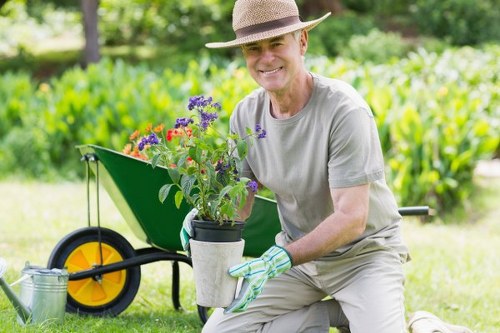A smiling mature man wearing a straw hat and light grey gardening clothes is kneeling on a lush green lawn in a well-maintained garden in Hendon, London. He is holding a black and grey plastic pot filled with vibrant, freshly-potted flowering plants, including purple, red, and orange blooms. Behind him, a yellow and black wheelbarrow with a green body and yellow wheel is visible, positioned on a paved pathway that runs through the garden. The background features neatly trimmed hedges, dense foliage, and tall trees, creating a peaceful outdoor setting perfect for gardening projects. The scene depicts daytime with natural sunlight illuminating the vibrant colors of the flowers and greenery, emphasizing an active gardening service environment offered by Landscapers Hendon, with a focus on flower bed creation and outdoor landscape enhancement in the Hendon area near postcode NW4. The overall composition highlights the cultivation and planting elements typical of professional garden maintenance and landscaping services, capturing the welcoming atmosphere of a well-kept outdoor space.