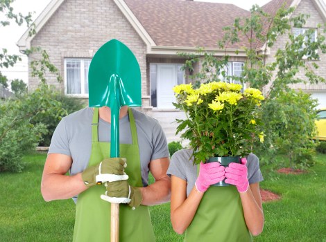 A person wearing a bright green gardening apron and yellow gloves is holding a garden fork upright, standing on a well-maintained lawn in front of a residential house with a grey tiled roof and cream-colored walls. Next to them, a second individual dressed similarly with pink gloves is holding a pink planter box filled with vibrant yellow chrysanthemums, partially obscuring their face. The garden features neatly trimmed grass, small landscaped shrubs, and a backdrop of young trees with light green foliage, suggesting a spring or early summer setting. The house in the background has white-framed windows and a central grey door, with a tidy front garden area and a wooden fence partially visible. The scene is evenly lit with natural daylight, indicating mild weather, and reflects a well-kept outdoor space suitable for gardening and landscaping services by Landscapers Hendon in the Hendon area, emphasizing outdoor maintenance and plant care.