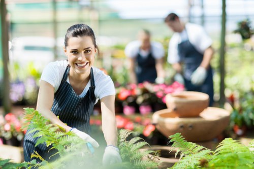 A smiling woman wearing a striped apron and gardening gloves is working in a well-lit greenhouse or garden centre, surrounded by lush green ferns and various flowering plants. Behind her, two other gardeners, slightly blurred, are tending to vibrant pink and red flowers on flower beds or display tables. The environment features glass panels allowing natural light to illuminate the outdoor setting, highlighting the healthy greenery and colorful blooms. The scene depicts an organized outdoor space or greenhouse area focused on plant care and gardening activities, with visible textures of soil, foliage, and concrete or stone pathways in the background. This image reflects professional gardening and landscaping work, consistent with services offered by companies like Landscapers Hendon, which serve the local area around Hendon, London, emphasizing quality plant care and outdoor maintenance.