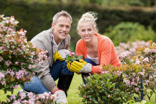 A cheerful man and woman are gardening together in a well-maintained outdoor space in Hendon, with lush green grass, blooming pink and purple flowering bushes, and a backdrop of trees and a hedge. The man has short gray hair and is wearing a light gray shirt, while the woman has blonde hair tied back and is dressed in a coral jacket and white top. They are both smiling while kneeling on the grass, wearing yellow gardening gloves, and holding a small potted plant between them. The garden features neatly trimmed shrubbery, a defined flower bed border, and appears to be part of a landscaped yard or front garden, reflecting professional outdoor maintenance and gardening services typical of Hedon or nearby areas. Overcast weather provides diffused natural light, highlighting the vibrant colours of the flowers and greenery, and emphasizing the welcoming, tidy environment suitable for garden landscape enhancements and flower bed ideas as promoted by landscapers Hendon.